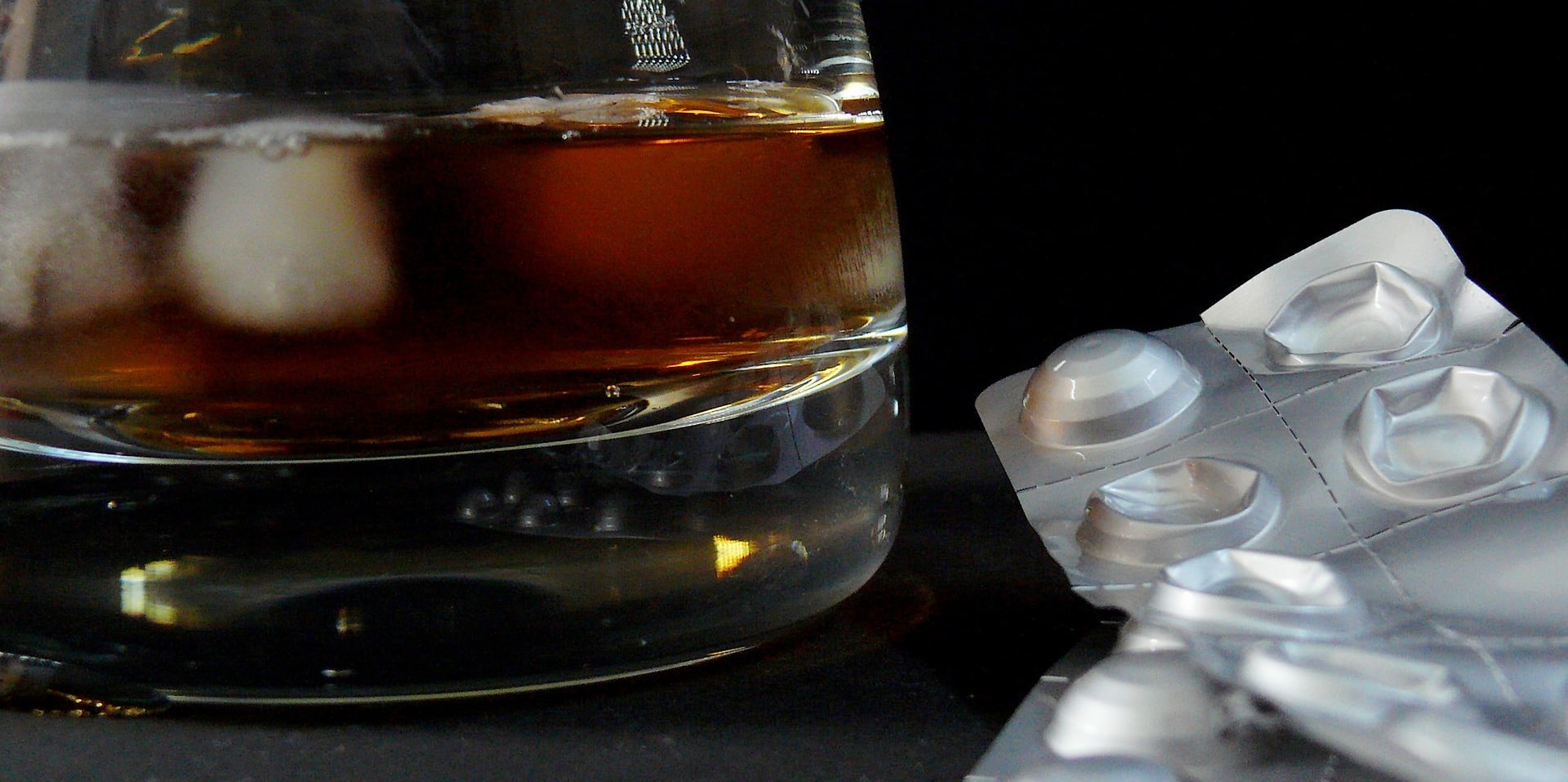 Glass tumbler of alcohol with ice sitting beside used pill packets