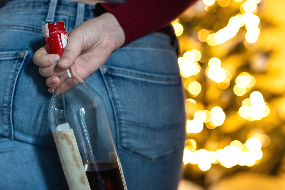 Woman concealing a wine bottle behind her near a Christmas tree with festive lights, representing secretive drinking tied to holiday stress.

